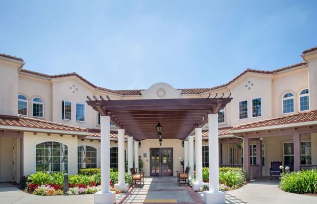 Main entrance of Sunrise at La Costa senior living facility with a shaded porch and landscaped area.
