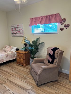 A cozy senior living bedroom featuring a comfortable chair, bed with floral bedding, and decorative wall elements.