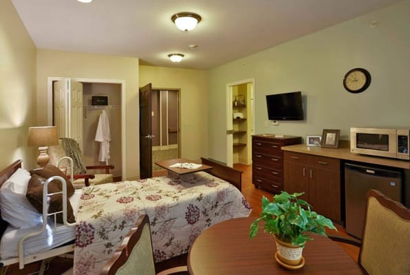 A cozy bedroom in a senior living facility with floral bedding, a TV, and wooden furniture.