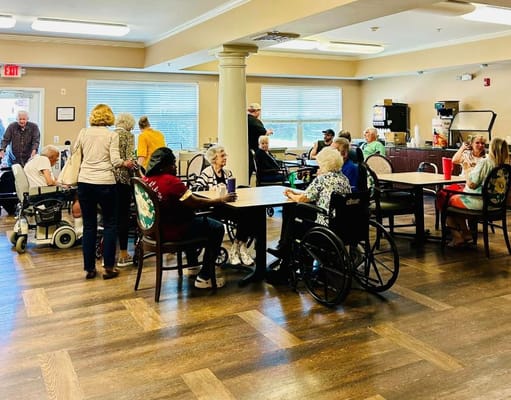Seniors socializing in a common area of a senior living facility, with some in wheelchairs.