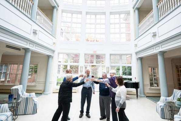 A group of six senior residents toasting with glasses in a bright atrium.