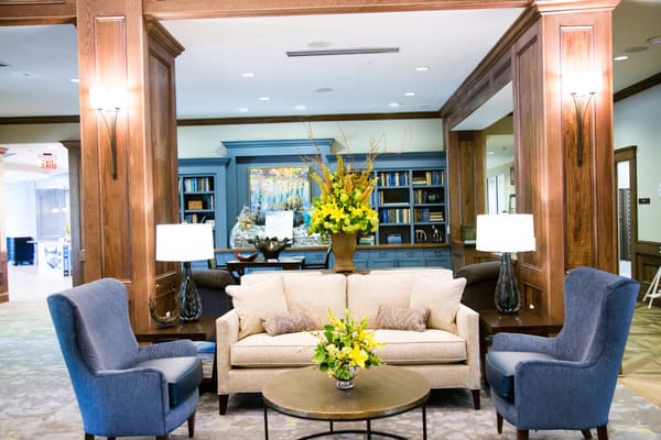 Cozy living room with two blue chairs and a beige sofa surrounded by bookshelves and floral arrangements.