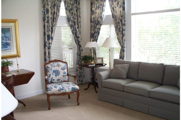 A living room featuring a floral armchair, a gray sofa, and large windows with decorative curtains.