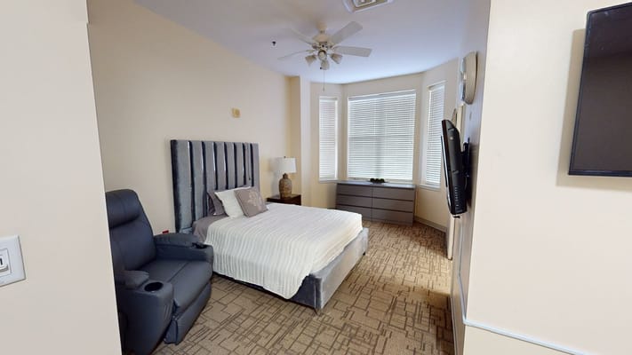 A cozy bedroom featuring a bed, armchair, dresser, and windows with blinds.