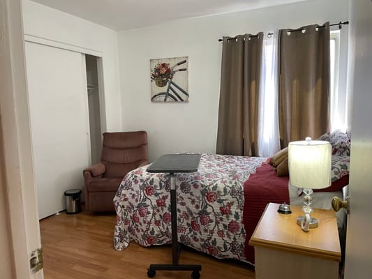 A cozy bedroom featuring a bed with floral bedding, a bedside table, a lamp, and a recliner chair.