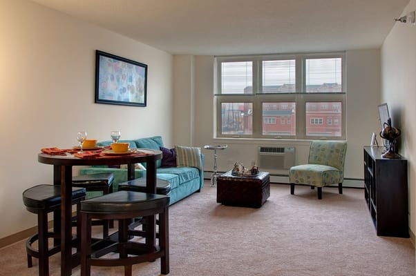 A living room featuring a teal couch, dining table, and large windows.