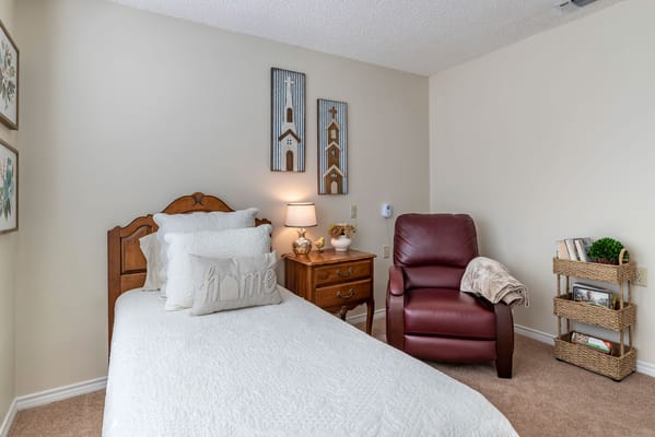 A cozy bedroom featuring a bed with white bedding, a side table with a lamp, and a comfortable chair.