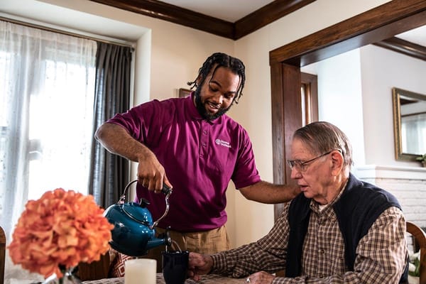 Caregiver pouring tea for a senior man at a dining table.