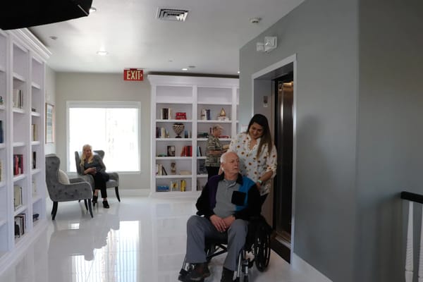 A caregiver assists a senior man in a wheelchair in a well-lit hallway, with two residents in the background.