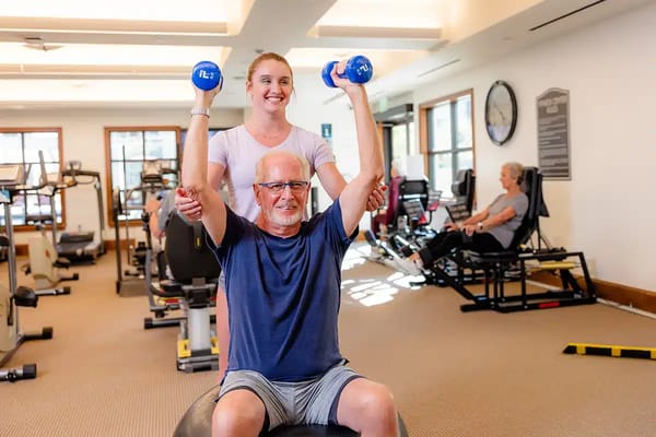 A senior exercising with weights, assisted by staff in a fitness room