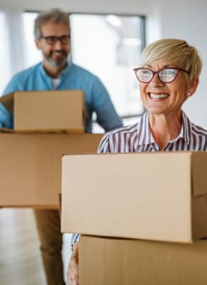 A cheerful older woman carrying boxes with a smiling man behind her