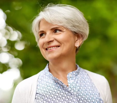 A smiling older woman outdoors with greenery in the background