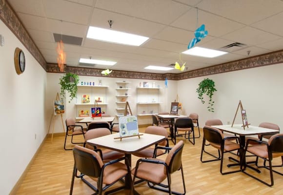 Indoor activity room with tables and chairs, decorated with plants and art displays.