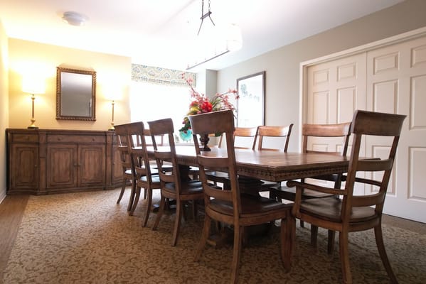 Brightly lit dining room with a large table and wooden chairs.