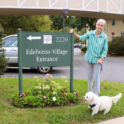 Senior woman standing by the Edelweiss Village entrance sign with a small dog.