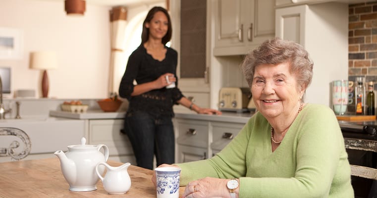 A senior smiling at a kitchen table with a caregiver in the background