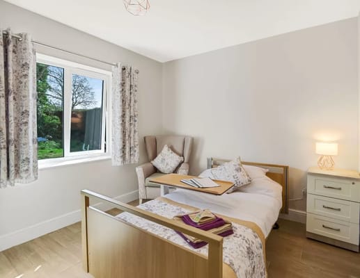 A well-furnished bedroom with natural light, featuring a bed, chair, and bedside table.