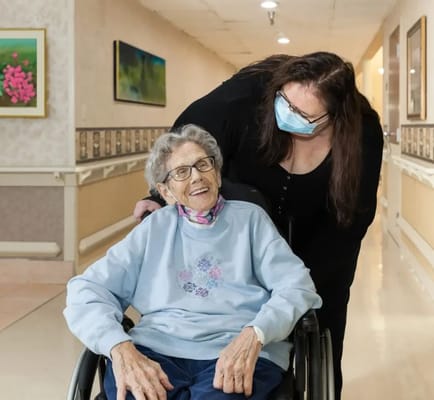 A smiling elderly woman in a wheelchair with a caregiver in a hallway.