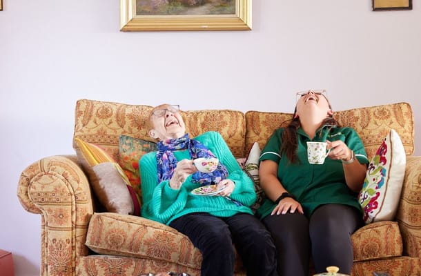 An elderly woman and a caregiver laughing while drinking tea on a sofa.