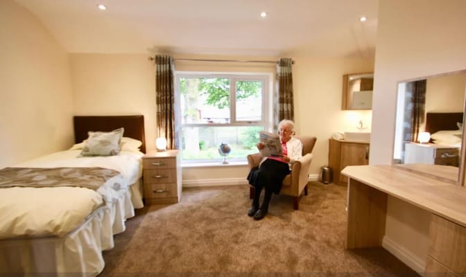 A senior woman reading a newspaper in a comfortable room with a bed and window.