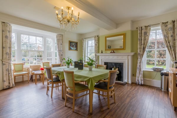 Bright dining room with a large table and chairs, featuring a chandelier and windows overlooking the garden.