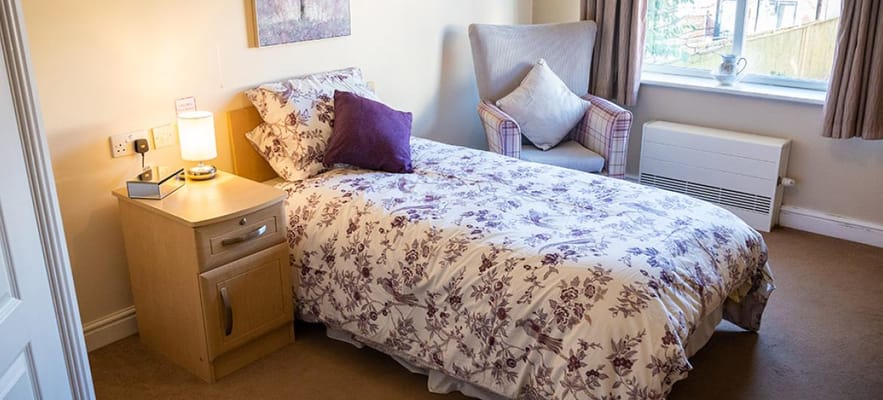 A cozy bedroom featuring a single bed with floral bedding, a bedside table with a lamp, and a comfortable armchair.