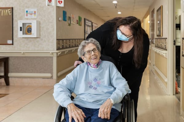 A staff member assisting a smiling resident in a wheelchair in a hallway.