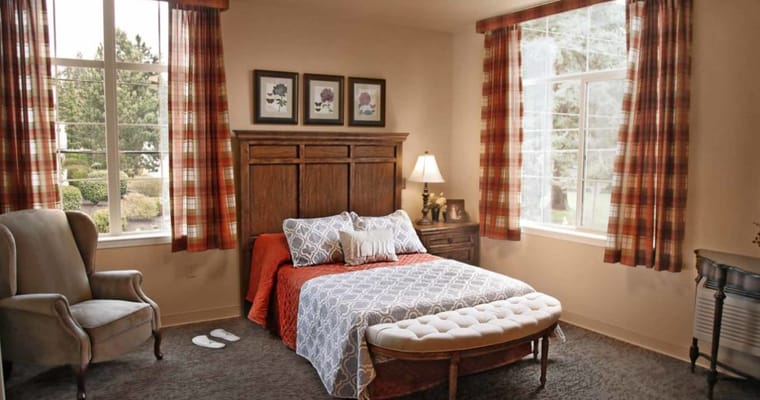 A cozy bedroom featuring a bed, armchair, and large windows with plaid curtains.