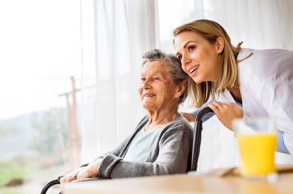 A caregiver smiles and leans in towards a senior woman in a bright room.