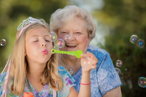 A girl blowing bubbles with an elderly woman smiling behind her.
