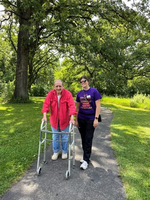 An elderly man using a walker with a caregiver in a park