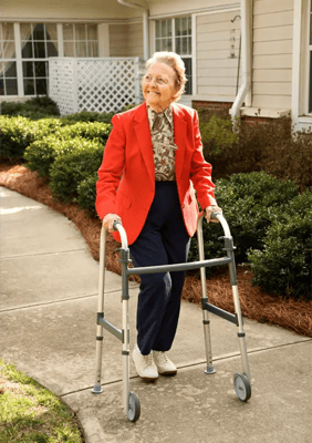An elderly woman using a walker on a paved path outdoors.