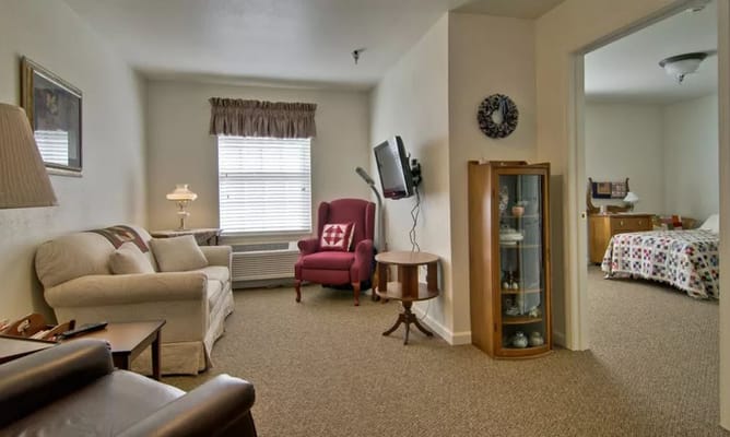 A cozy living room in a senior living facility with a sofa, armchair, and a view into a bedroom.