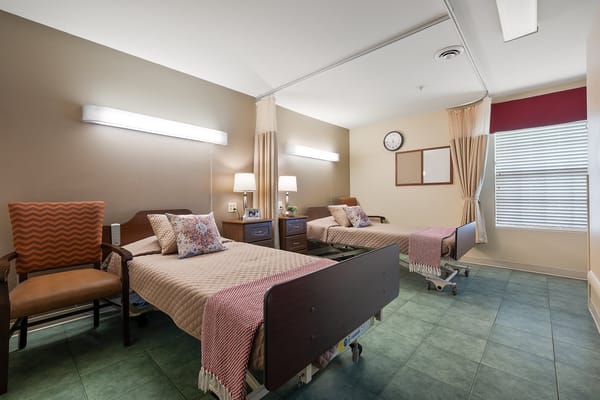 A shared bedroom in a senior living facility with two beds and cozy furnishings.