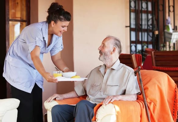 Caregiver serving food to an elderly man in a cozy living area