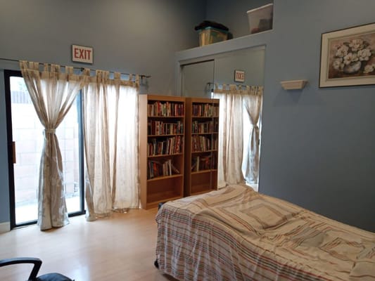 A well-lit room with a bed, bookshelf, and large windows with curtains.