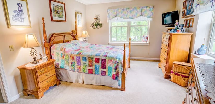 A well-lit bedroom featuring a bed with a colorful quilt, wooden furniture, and a window.