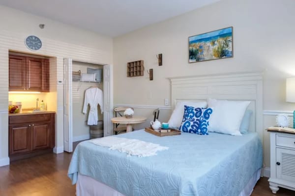 A well-decorated bedroom featuring a bed with a blue cover, kitchenette, and cozy sitting area.