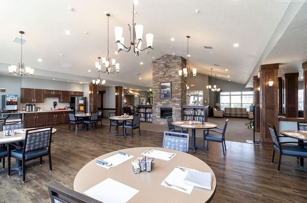Spacious dining area with tables and chairs, featuring a stone fireplace and bookshelves.