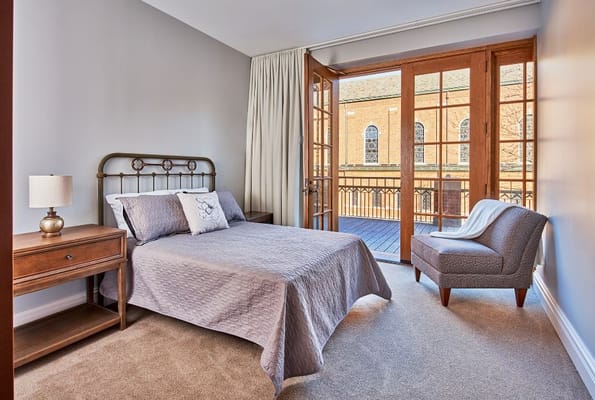 A cozy bedroom with a bed, lamp, and armchair, featuring French doors that open to a balcony.