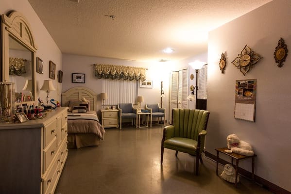 A well-furnished bedroom with a bed, dresser, and seating area in Jennifer Gardens Assisted Living.