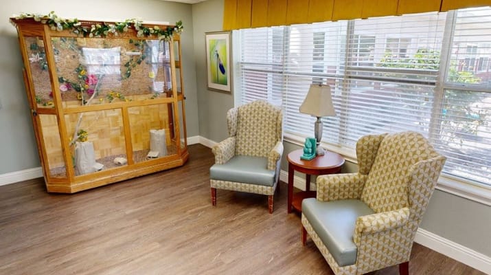 A cozy living room with two armchairs, a side table, and a display case in a senior living facility.