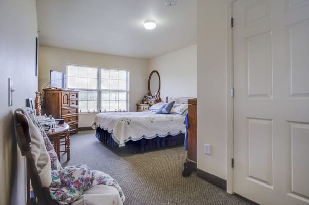 A cozy bedroom with a bed, dresser, and chair, featuring large windows.