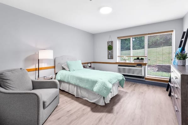 A cozy bedroom featuring a bed with a light blue quilt, armchair, and window with natural light.
