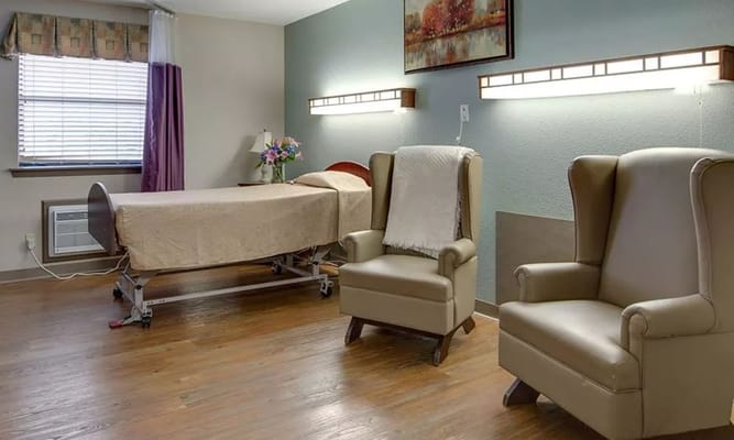 A cozy resident room featuring a bed, two chairs, and natural light.