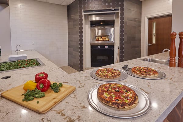 Three pizzas on metal trays in a kitchen with fresh vegetables on a cutting board