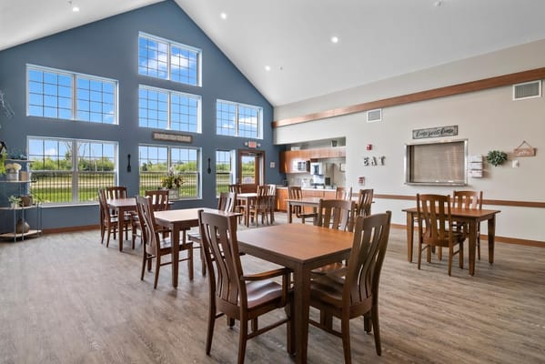 Bright dining area with wooden tables and chairs, large windows, and kitchen space.