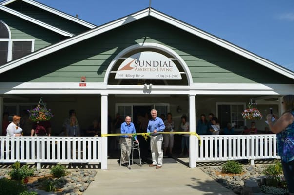 Two men cutting a ribbon at the entrance of Sundial Assisted Living with a crowd in attendance.