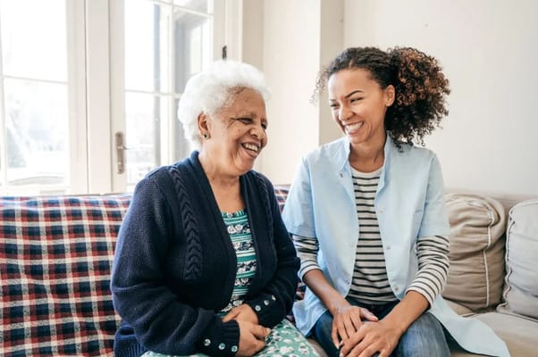 A senior woman and a caregiver laughing together on a couch.