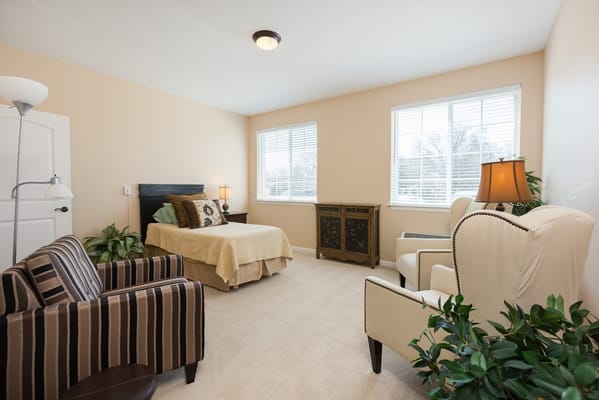 A bedroom with a bed, two chairs, and large windows at Three Oaks Assisted Living.